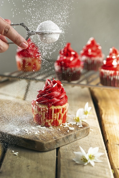 Vertical shot of a person adding sugar powder on a cupcake with red cream through a strainer