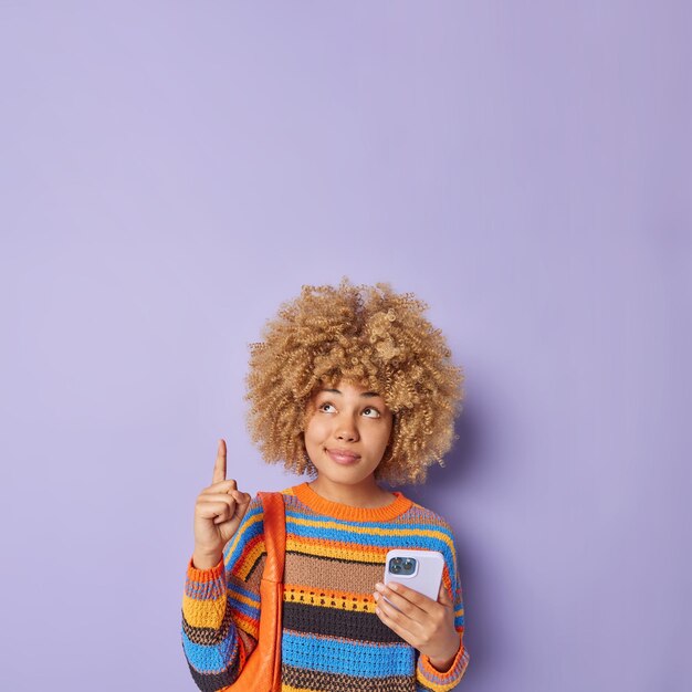 Vertical shot of pleasant looking curly haired young woman
points index finger overhead holds modern cellular demonstrates
blank space for your advertising content isolated over purple
background