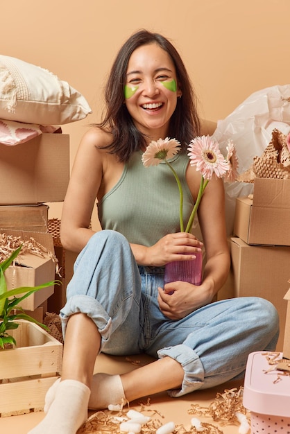 Vertical shot of positive asian woman dressed casually holds\
vase with gerbera flowers applies green hydrogel patches under eyes\
to remove wrinkles relocates to new apartment surrounded by\
boxes