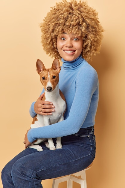 Vertical shot of positive curly woman sits on chair with\
favorite dog poses im studio for memorable photo wears blue\
turtleneck and jeans isolated over beige background people animals\
and friendship