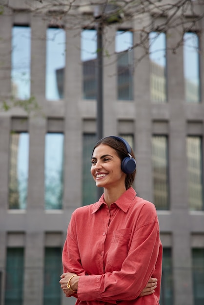 Vertical shot of positive dark haired millennial girl listens music in headphones while strolling in city wears red shirt keeps arms crossed looks happily away poses near modern building outdoors