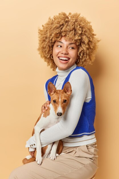 Vertical shot of positive woman with curly hair dressed in\
casual clothes poses with pedigree dog sits on chair enjoys company\
of pet isolated over beige background people animals and\
friendship