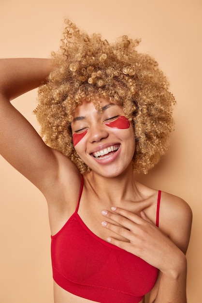 L'immagine verticale di una giovane donna positiva con i capelli ricci indossa un top corto rosso applica cerotti idrogel sotto gli occhi per il trattamento della pelle sorrisi ampiamente isolati su sfondo beige concetto di bellezza