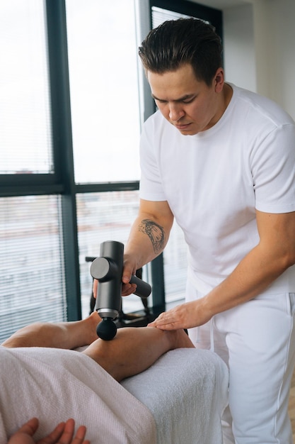 Vertical shot of professional male masseur massaging leg calf muscles using massage gun percussion tool of muscular athlete man on spa treatment lying on back in massage table