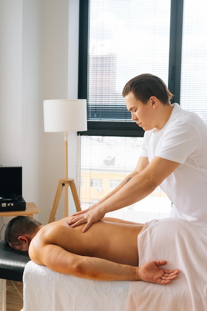 Vertical shot of professional male masseur with strong tattooed\
hands massaging back and shoulder of muscular sports man lying on\
stomach