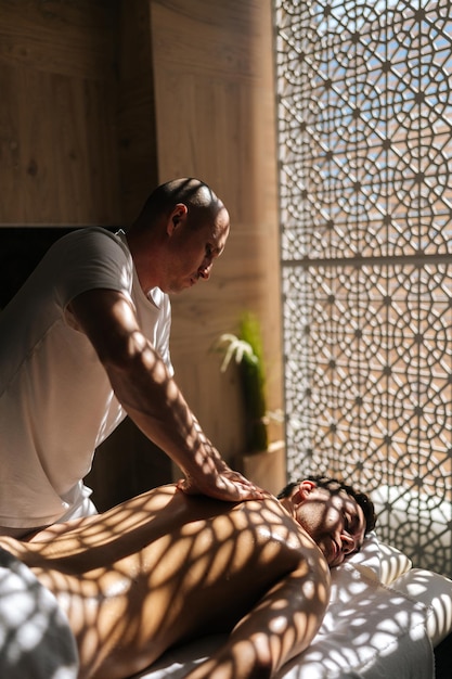 Photo vertical shot of professional masseur male doing back spa massage of man in massage room lying on table in sunlight and shadows in spa center relaxed client came to massage therapy due to body aches