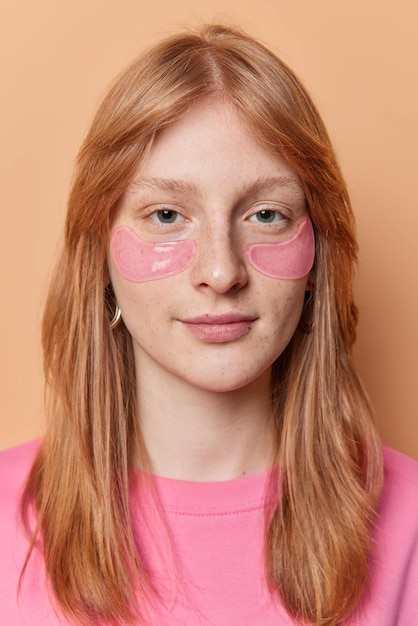 Vertical shot of serious pretty redhead girl applies hydrogel patches under eyes reduces bags looks directly at camera poses against brown background has clean freckled skin healthy complexion