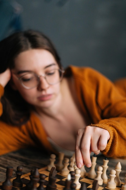 Vertical shot of serious sexy young woman making chess move lying on wooden vintage floor in dark room