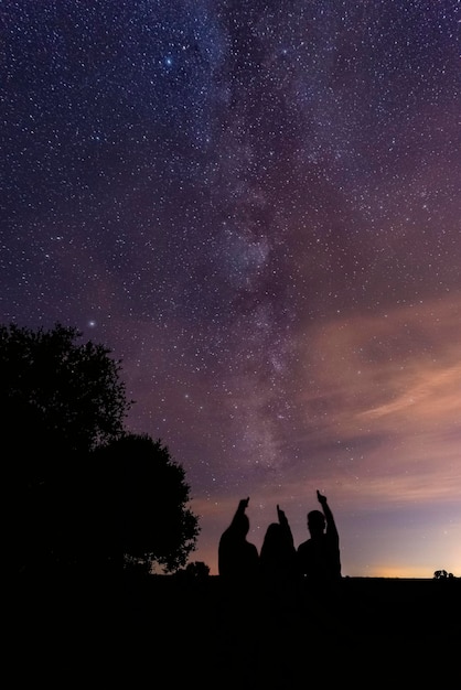 Вертикальный снимок силуэтов трех человек, указывающих на млечный путь в ночном небе