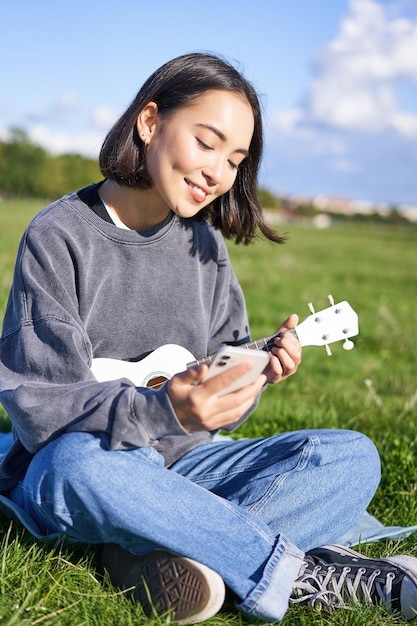 스마트폰으로 미소 짓는 아시아 소녀가 우쿨레를 연주하고 악보나 가사를 읽는 수직 