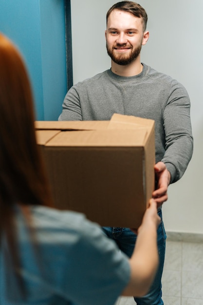 Vertical shot of smiling bearded courier male delivering cardboard box parcel to female customer on doorway at home