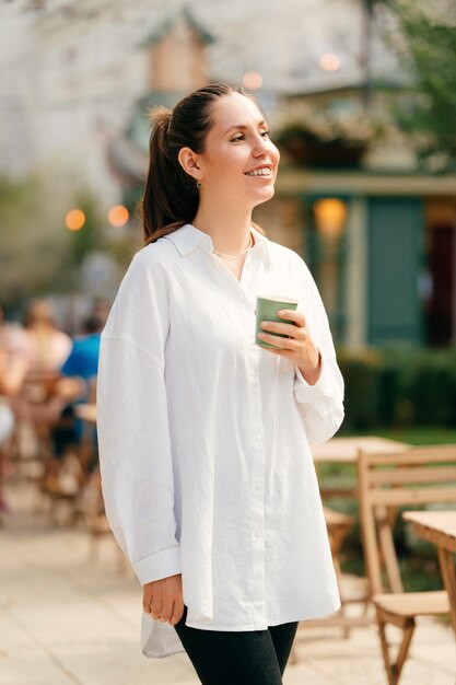 Foto colpo verticale di una donna sorridente e sicura che si gode il caffè mattutino per le strade della città