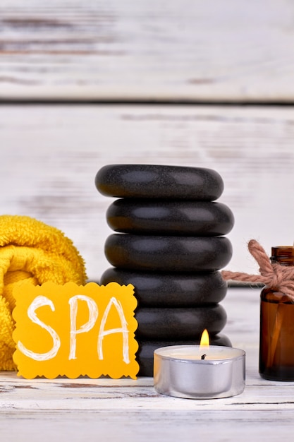 Vertical shot spa treatment accessories on white wooden background. Stacl of black polished stones with candle and rolled towel.