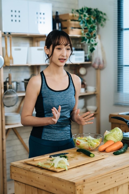 집에 있는 부엌에서 온라인으로 튜토리얼 비디오를 만드는 샐러드를 촬영하는 동안 손 제스처를 사용하는 낚시를 좋아하는 아시아 여성 인플루언서의 수직 샷.