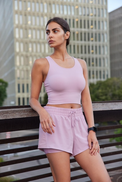 Colpo verticale di una giovane donna sportiva vestita con top corto e pantaloncini che riposa dopo aver fatto esercizi sportivi distoglie lo sguardo in posa sul ponte sullo sfondo urbano. la modella femminile attiva ha un allenamento mattutino