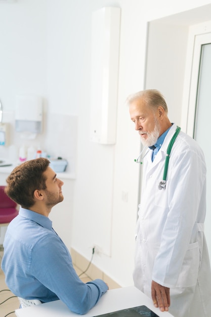 Vertical shot of standing mature male adult doctor examining male patient sitting at table in medical office. Handsome young man client listening to older senior physician explaining treatment.