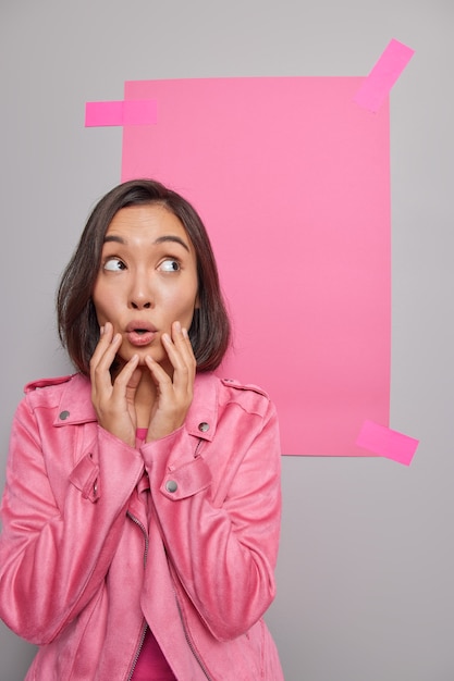 Vertical shot of stunned brunette Asian woman keeps hands on face dressed in pink jacket reacts on stunning news focused above poses against studio wall