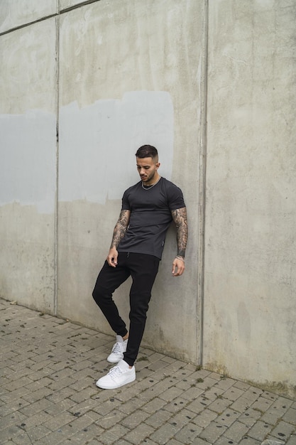 Vertical shot of a stylish European man with tattooed arms leaning on a wall