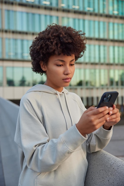 Vertical shot of thoughtful curly haired woman searches\
internet on smartphone dressed in casual hoodie checks newsfeed in\
social networks poses against blurred city building has break on\
workout