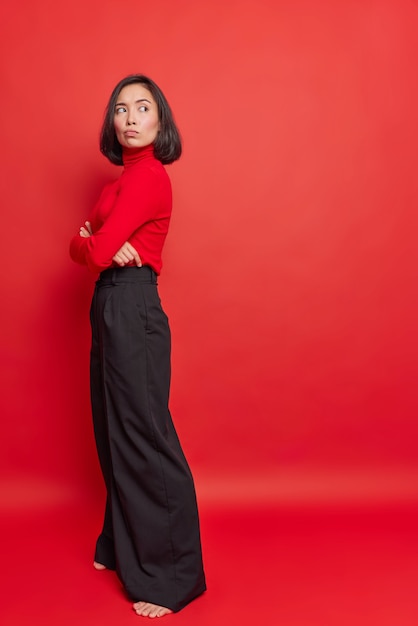 Vertical shot of thoughtful displeased Asian woman with dark hair keeps arms crossed turns away being dissatisfied with something wears turtleneck and black trousers stands against red wall