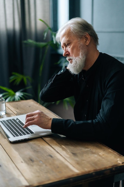 Vertical shot of tired gray-haired mature business man sitting\
at workplace experiences severe pain in neck, rubbing it to relieve\
muscle tension. senior man suffering from neck pain at table with\
pc.