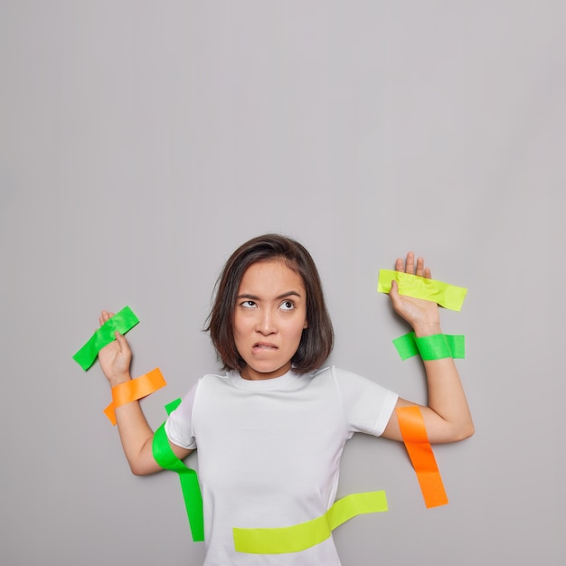 Vertical shot of unhappy Asian woman bites lips looks angrily above being captured plastered to wall with adhesive tapes isolated over grey wall