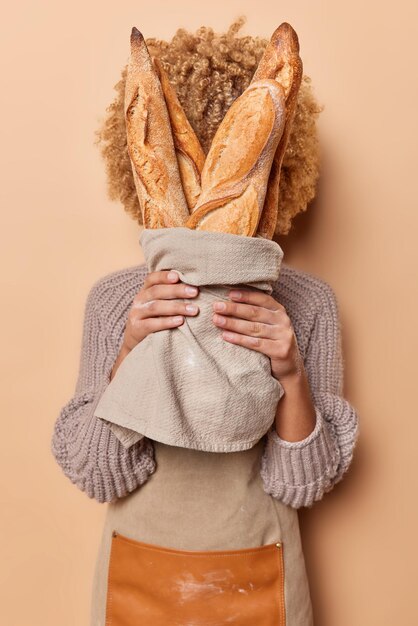Vertical shot of unknown curly haired woman covers face with\
fresh baked baguettes wrapped in linen napkin wears knitted jumper\
and apron isolated over beige background bread baking concept