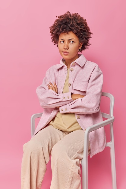 Vertical shot of upset offended young woman with curly hair keeps arms folded dressed in stylish clothes poses on comfortable chair isolated over pink wall