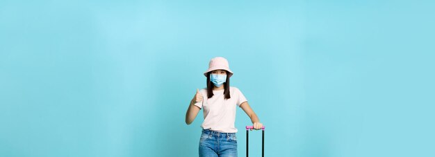 Vertical shot of vaccinated asian girl in medical face mask going on vacation standing with suitcase