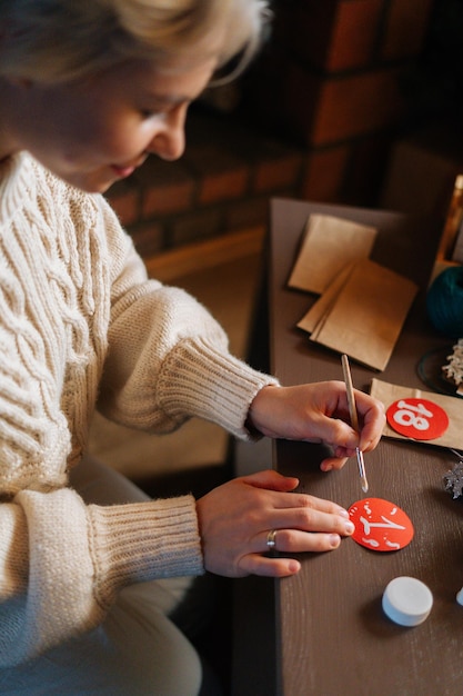 Photo vertical shot of young woman writing number on red bag making paper bags from kraft paper for advent