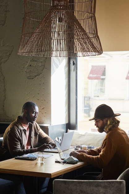 Foto vista laterale verticale ritratto di due uomini contemporanei che utilizzano computer portatili mentre lavorano al tavolo del bar illuminato dalla luce del sole nell'interno del loft, spazio copia