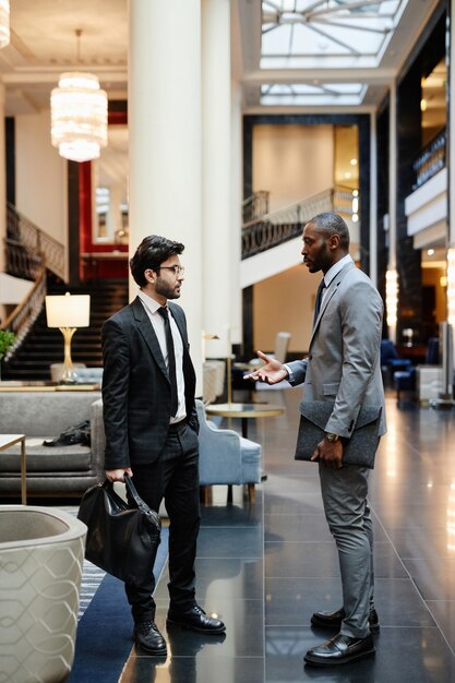 Vertical side view portrait of two successful businessmen discussing work while standing in hotel lobby
