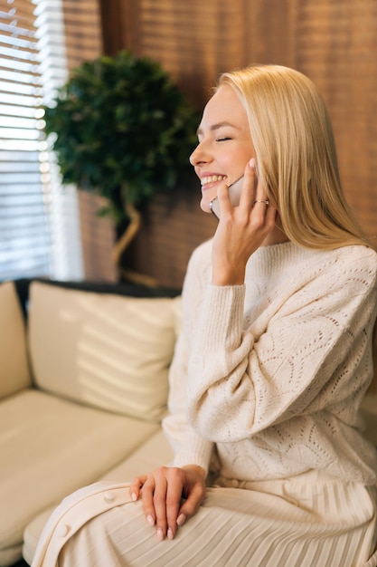 Vertical side view shot of joyful young woman talking on mobile phone sitting alone on sofa by window in cafe closed eyes Happy blonde female conversation with smartphone while rest during breakfast