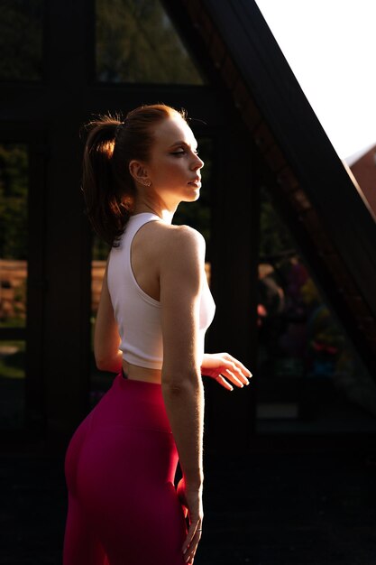 Vertical side view of sporty female athlete preforming fitness training session at country house building exterior Healthy woman warming up outdoors on summer