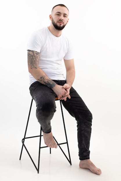 Vertical of tattooed handsome darkhaired confident bearded barefoot man sit on chair fashionable mod