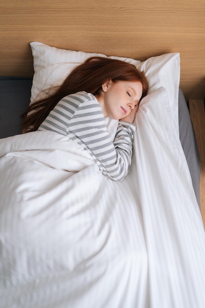 Vertical top view of cute redhead young woman sleeping well in bed hugging soft white blanket at home. Charming lady resting enjoying fresh soft bedding linen and mattress in bedroom.