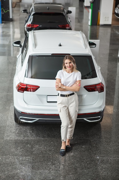 Vertical top view shot of a happy woman posing near her new automobile at car dealership