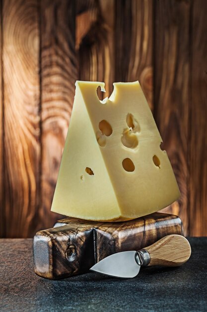 Вертикальный вид большой кусок молочного сыра маасдам на маленькой разделочной доске и нож на старом деревянном фоне