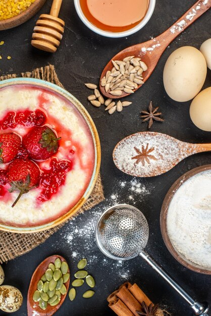 Vista verticale della deliziosa colazione servita con marmellata di fragole in una ciotola e miele, cannella, lime, uova, cucchiai, su, colore scuro, fondo