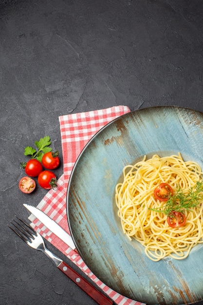 Vertical view of delicious spagetti served with tomatoes greens and cutlery set on red stripped towel on the left side on black background