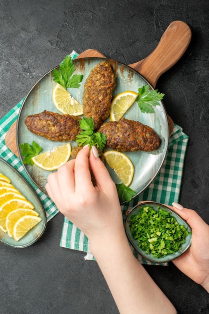 Vertical view of hand putting green on delicious cutlets served with lemon and green on wooden cutting board on yellow stripped towel on black background