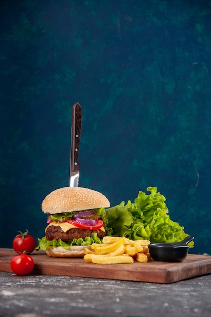 Vertical view of knife in meat sandwich and fries tomatoes with stem on wooden board ketchup on dark blue surface