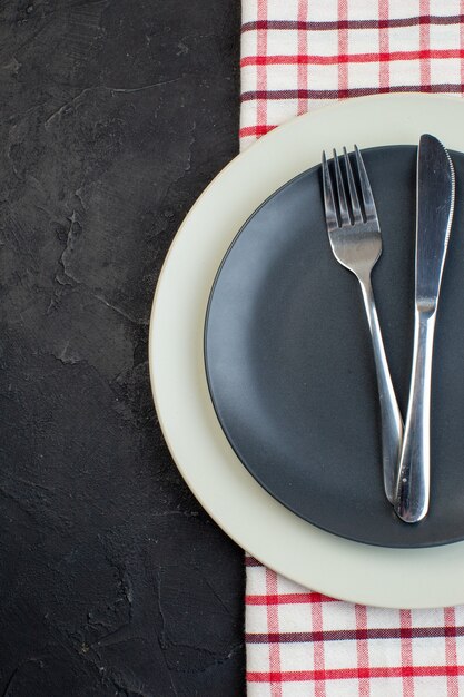 Vertical view of stainless cutlery set on dark gray color and white empty plates on red stripped towel on the left side on black background with free space