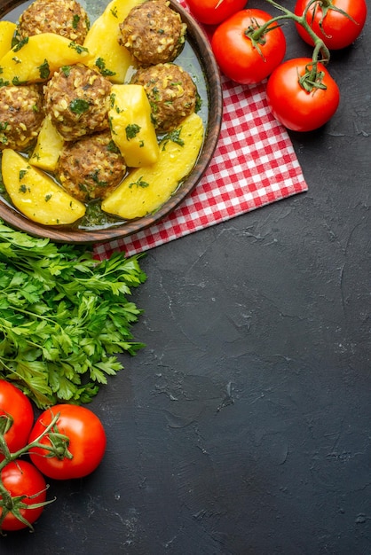 Vertical view of tasty dinner with meatballs potatoes green in a brown bowl on red stripped towel tomatoes on black background with free space