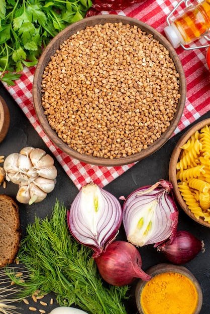 Vertical view of uncooked buckwheat in a brown bowl on towel pasta in pot fresh vegetables yellow ginger green bundle bread slices