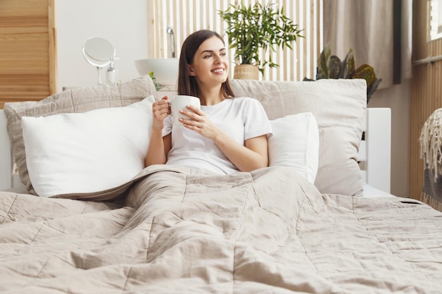 Vertical view of woman sitting at the bed with a cup of coffee and looking to the window