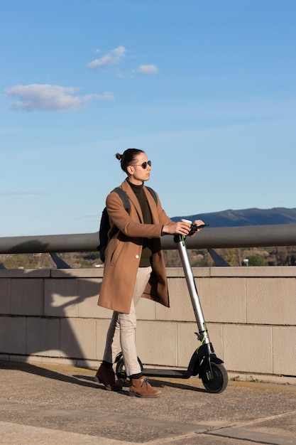 Vertical view of young handsome man with bun hairsyle walking with his electric scooter as and alternative transport