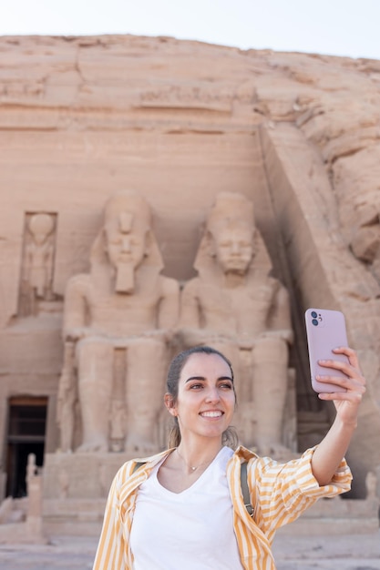 Photo vertical view of young smiling tourist woman taking a selfie in the facade entrance of the abu simbel temple in egypt