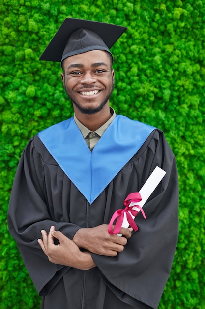 Vertical waist up portrait of happy African-American man wearing graduation gown and smiling at camera while standing against green plant background