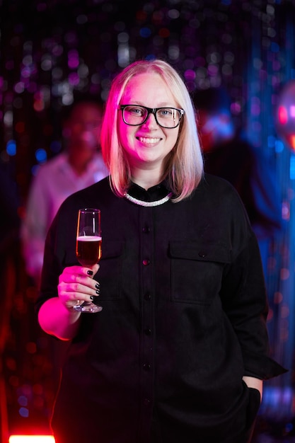 Vertical waist up portrait of smiling woman holding drink at\
party in neon light